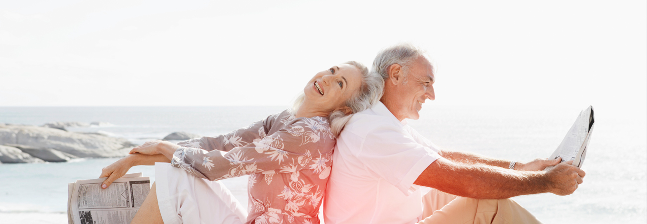 Älteres Paar sitzt lachend Rücken an Rücken am Strand