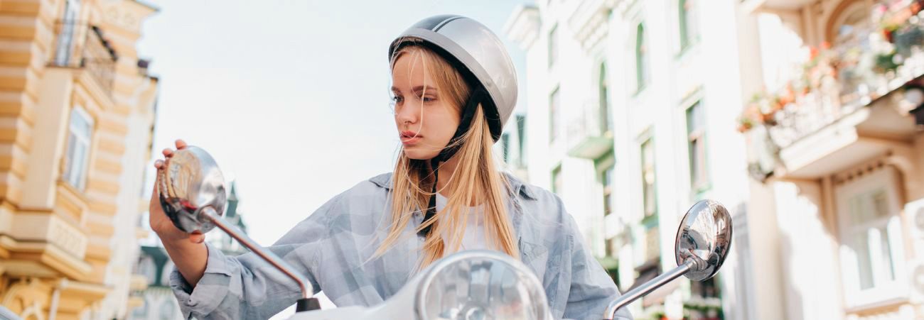 Junges blondes Mädchen mit Helm sitzt auf einem Roller und stellt die Spiegel ein