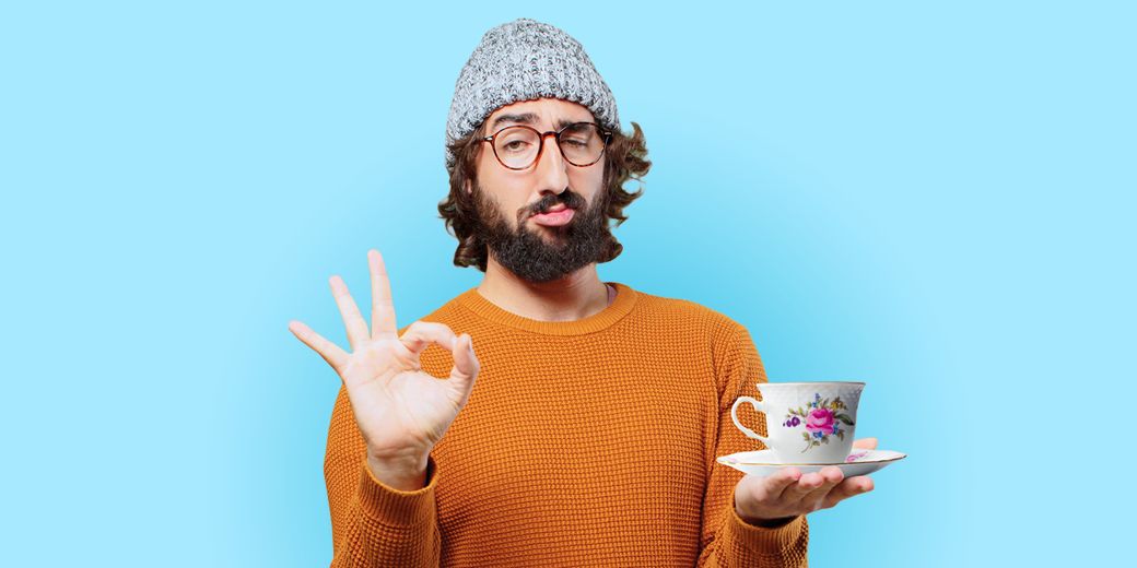 Bärtiger Hipster mit Brille und Mütze hält Teetasse aus Porzellan und zeigt Okay-Geste als Symbol für Hausratversicherung und Schutz wertvoller Dinge im Zuhause.