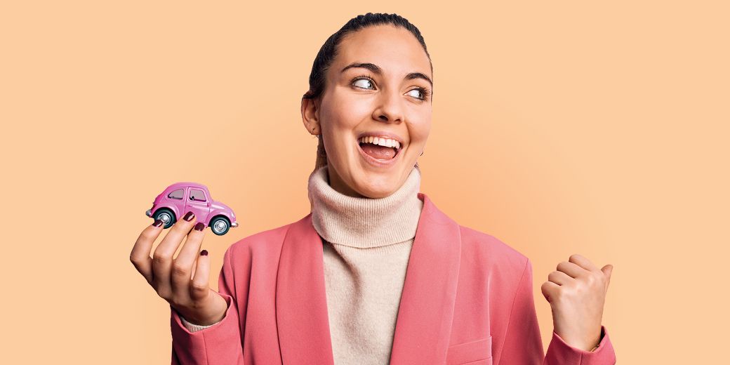 Junge Frau in rosa Blazer hält ein kleines Spielzeugauto in der Hand und freut sich 