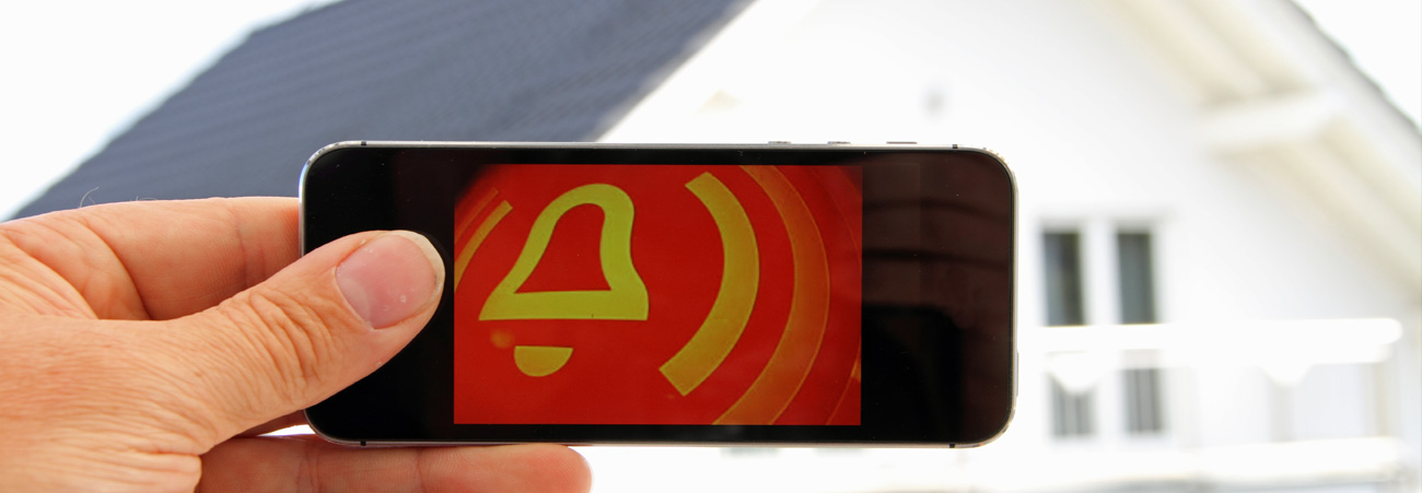 Hand hält Smartphone mit rot-gelben Alarmsignal auf dem Display, im Hintergrund ein Haus