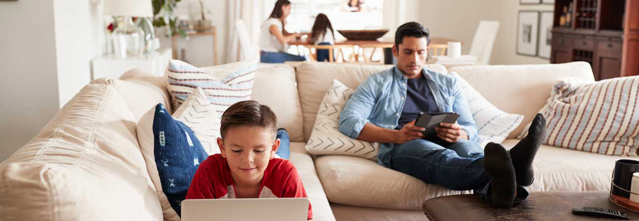 Familie im Wohnzimmer: Kind nutzt Laptop auf dem Sofa, Vater entspannt daneben mit Tablet, Mutter arbeitet im Hintergrund am Tisch
