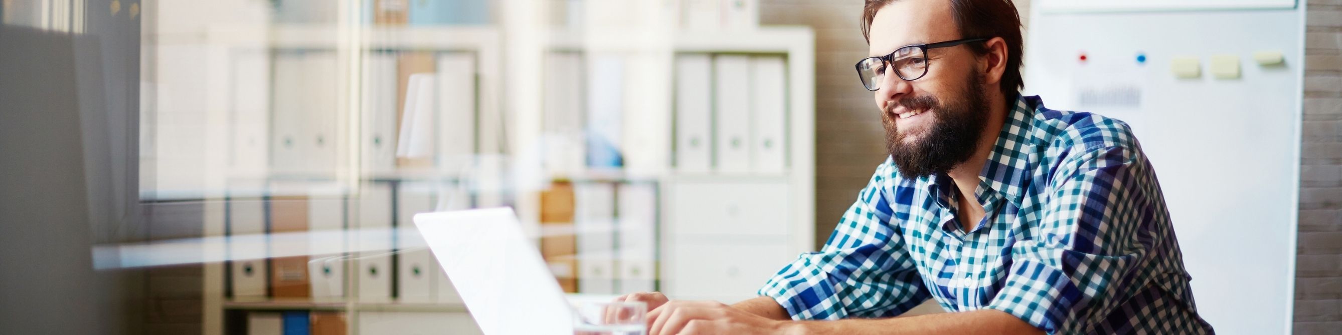Mann mit Brille arbeitet lächelnd am Laptop in einem modernen Büro