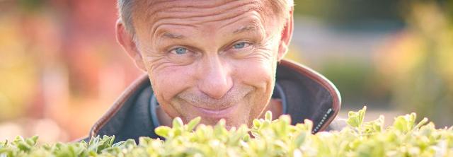 Älterer Mann schaut lächelnd über eine Gartenhecke