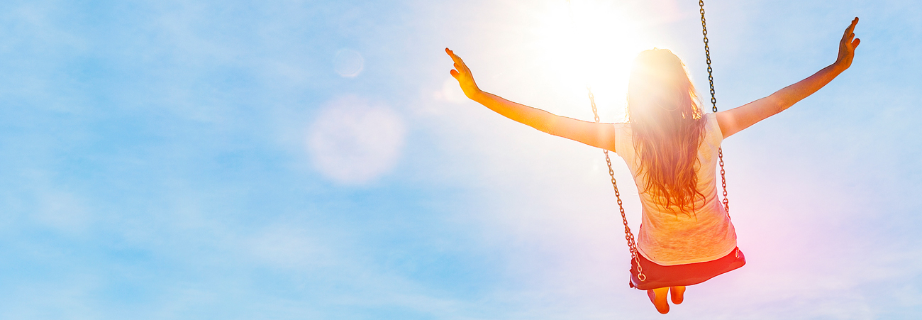 Frau sitzt auf Schaukel und schaut in den blauen Himmel und die Sonne