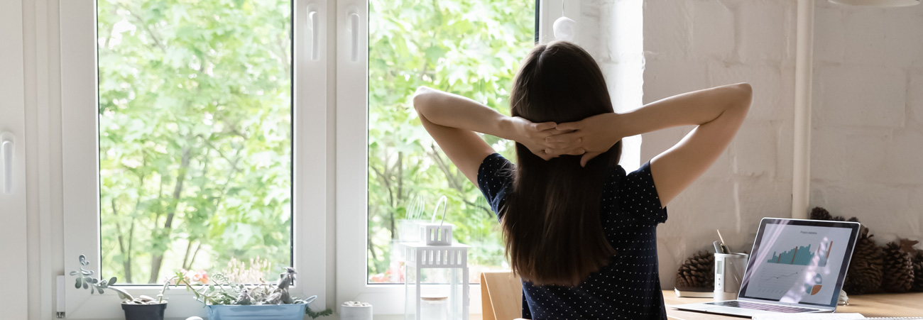 Junge Frau sitzt mit hinter dem Kopf verschränkten Armen am Schreibtisch.