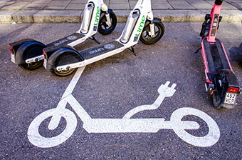 E-Scooter stehen am Straßenrand, auf dem Asphalt eine weiße Markierung eines E-Scooters mit Stecker-Symbol