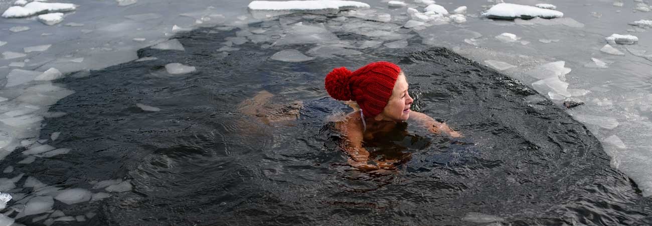 Person mit roter Mütze beim Eisbaden in einem zugefrorenen See, umgeben von Schnee und Eis