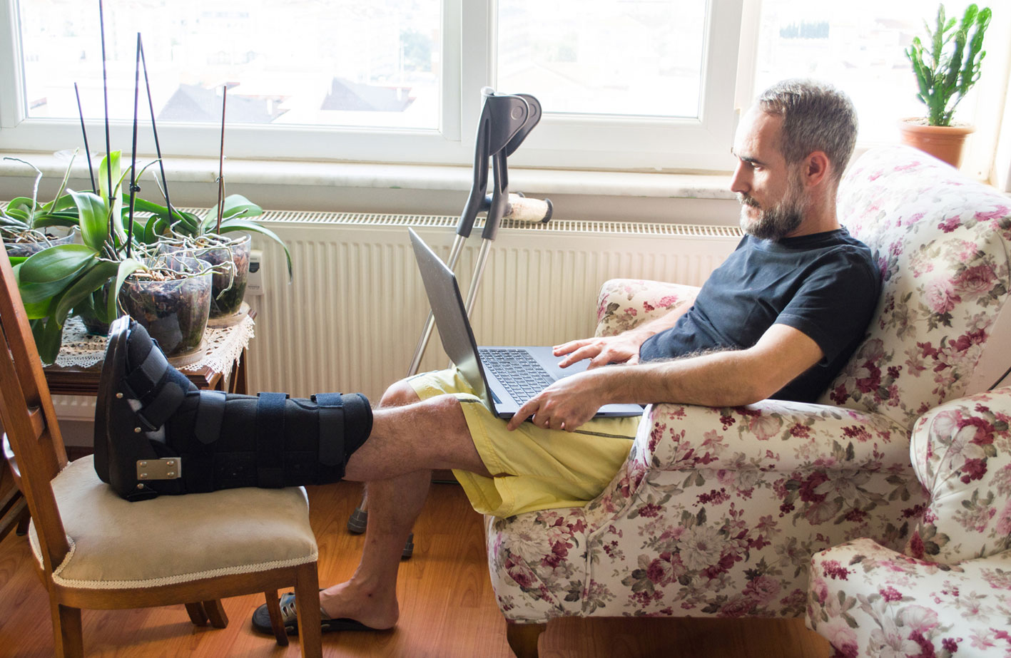 Mann mit Fußverletzung sitzt mit Laptop zu Hause auf dem Sofa