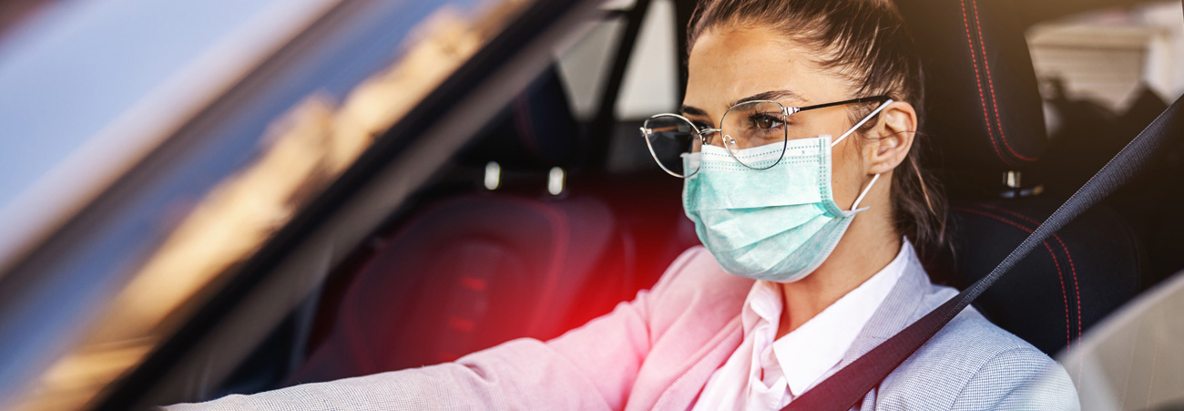 Junge Frau mit Brille und Mund-Nasen-Schutz sitzt am Steuer eines Autos.