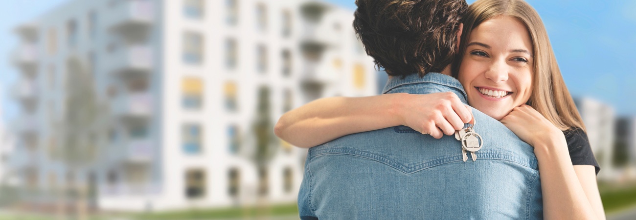 Junges Paar umarmt sich freudig mit Schlüssel in der Hand, Hintergrund: Häuserblock