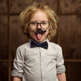 Lachendes Kind mit Brille, falschem Schnurrbart und Fliege streckt spielerisch die Zunge heraus