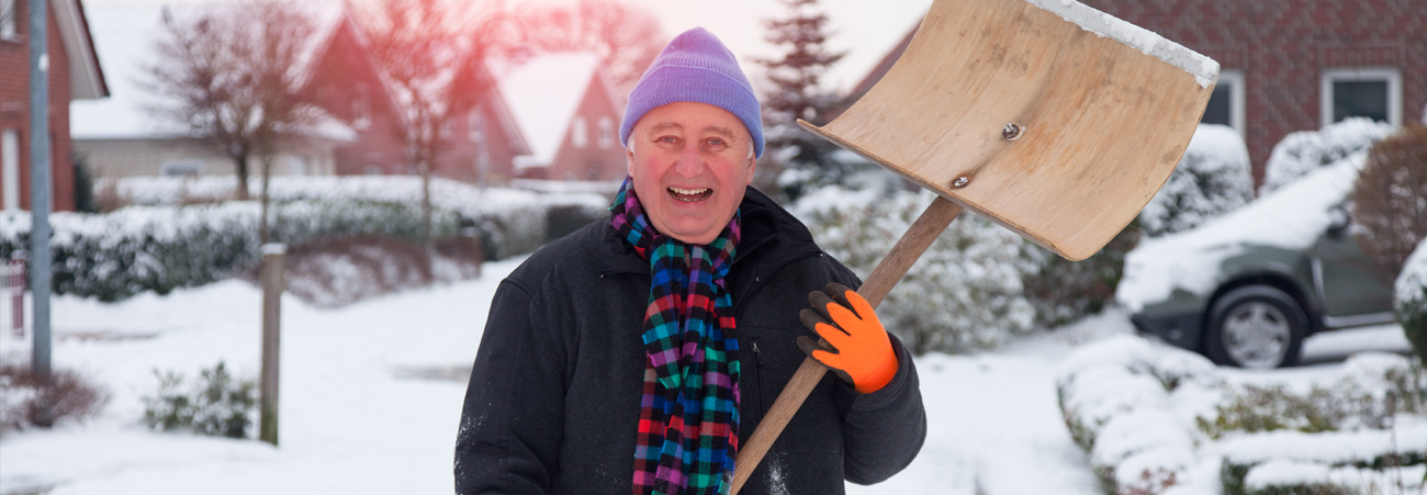 Älterer Herr steht lächelnd mit Schneeschieber in der Hand vor seinem Haus