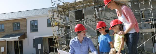 Vierköpfige Familie mit Bauhelmen steht vor dem Rohbau ihres Hauses